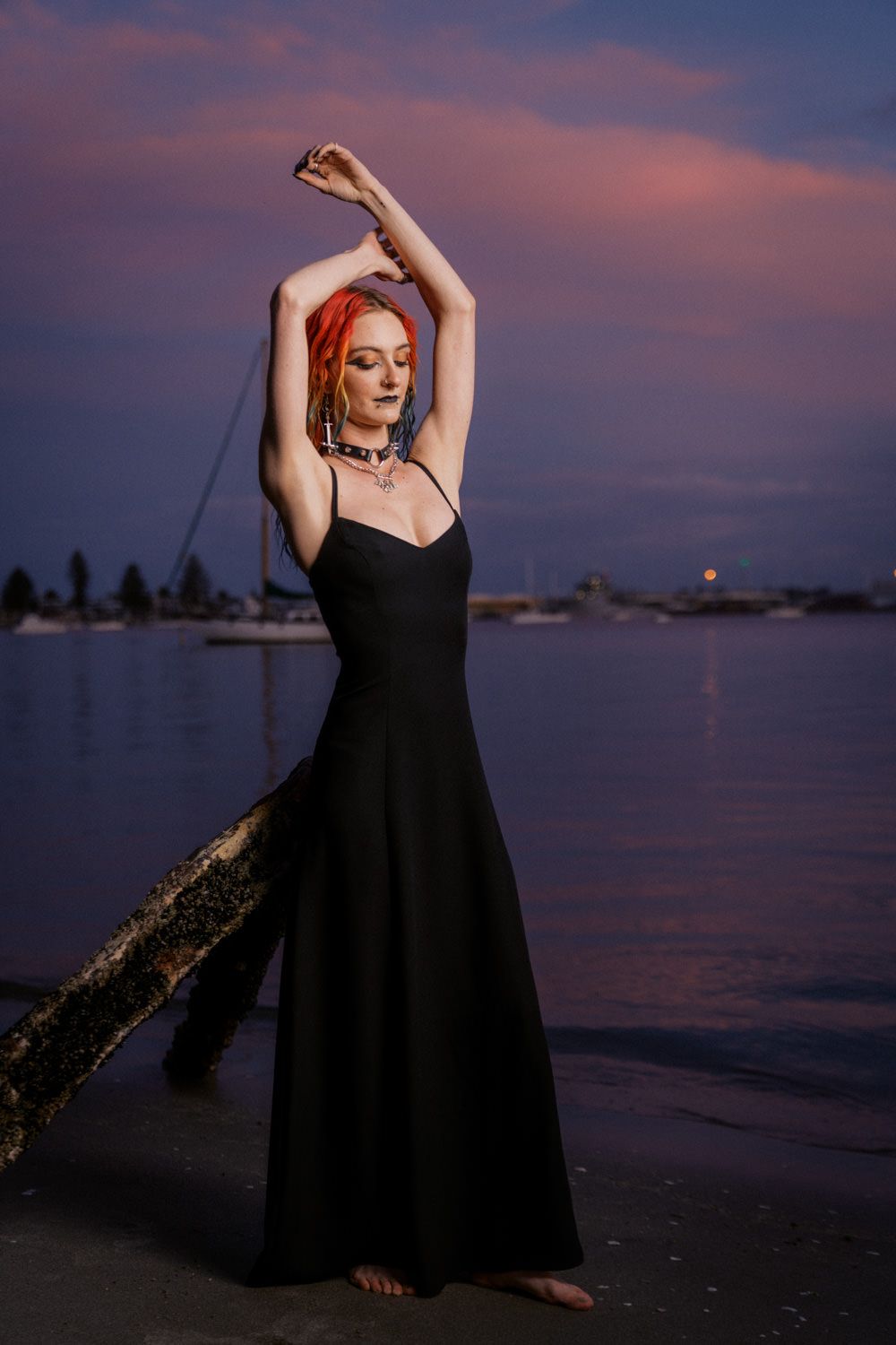 Woman in black gown with red hair poses gracefully on beach at sunset with purple sky.