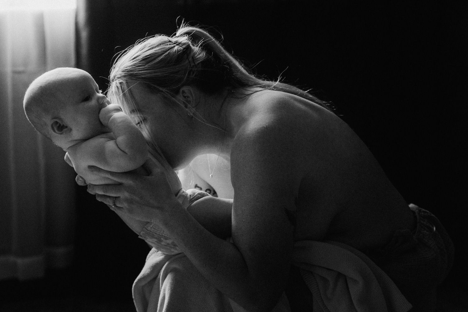 Black and white photo of a mother tenderly kissing her newborn baby in a softly lit room.