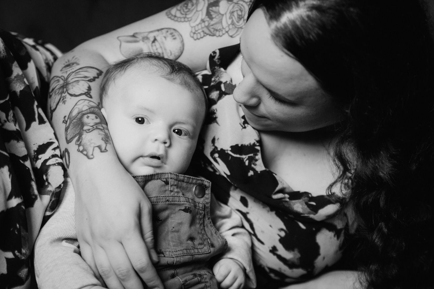 Black and white photo of a tattooed mother tenderly kissing her newborn baby while cradling them close.