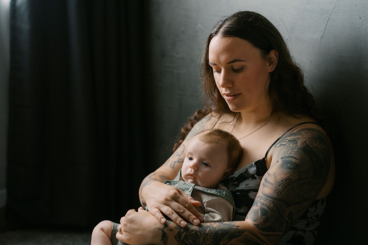 Tattooed mother tenderly holding her newborn baby against a dark background.