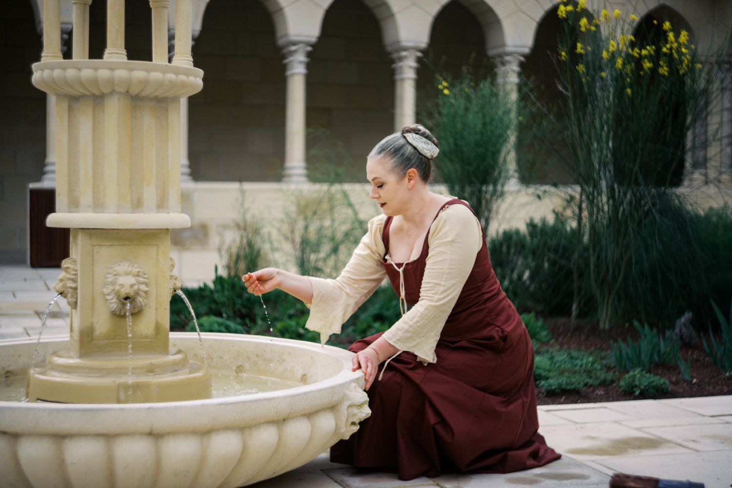Woman in medieval dress reaching toward ornate stone fountain in courtyard setting.