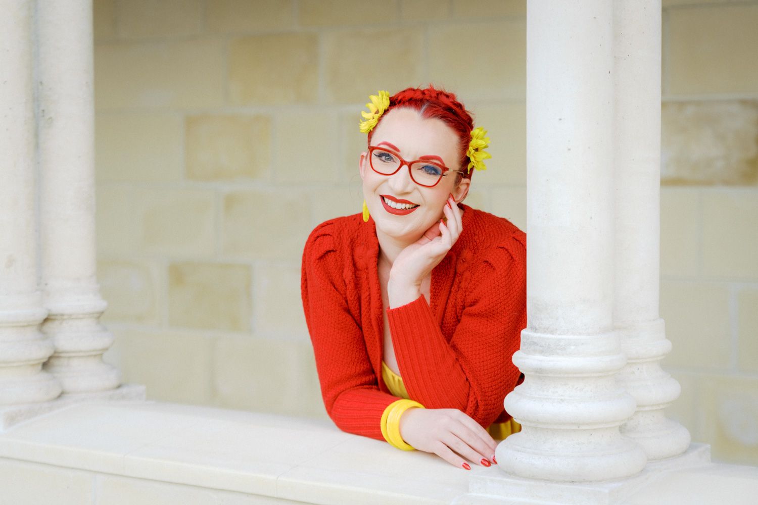 Woman with red hair and glasses wearing a red sweater posing gracefully between white columns outdoors.