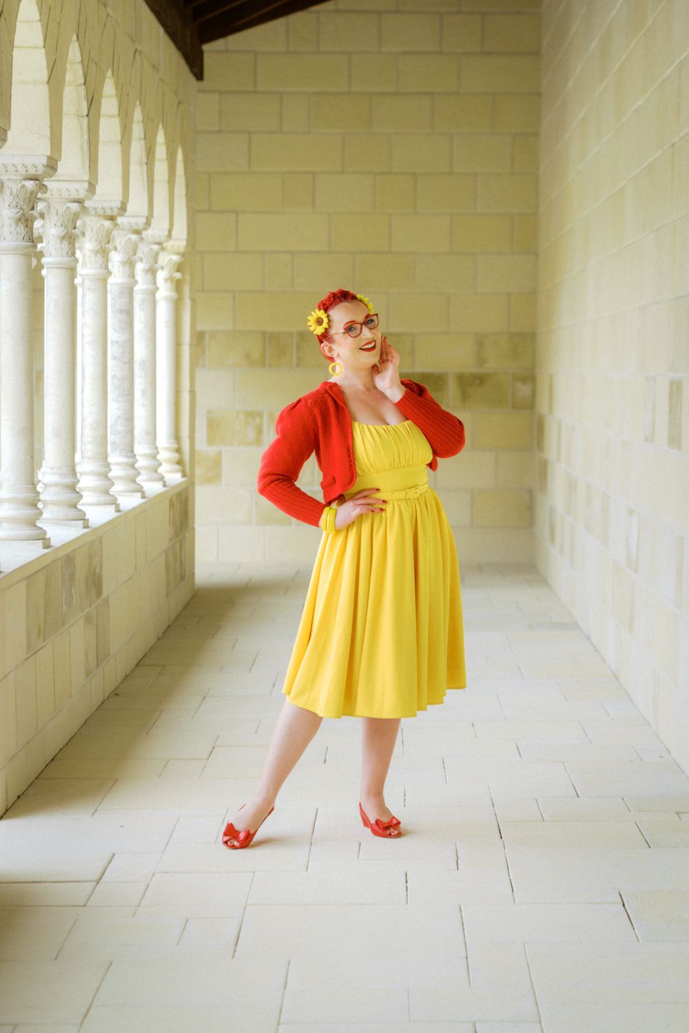 Woman in yellow dress and red cardigan poses in elegant white stone colonnade corridor.
