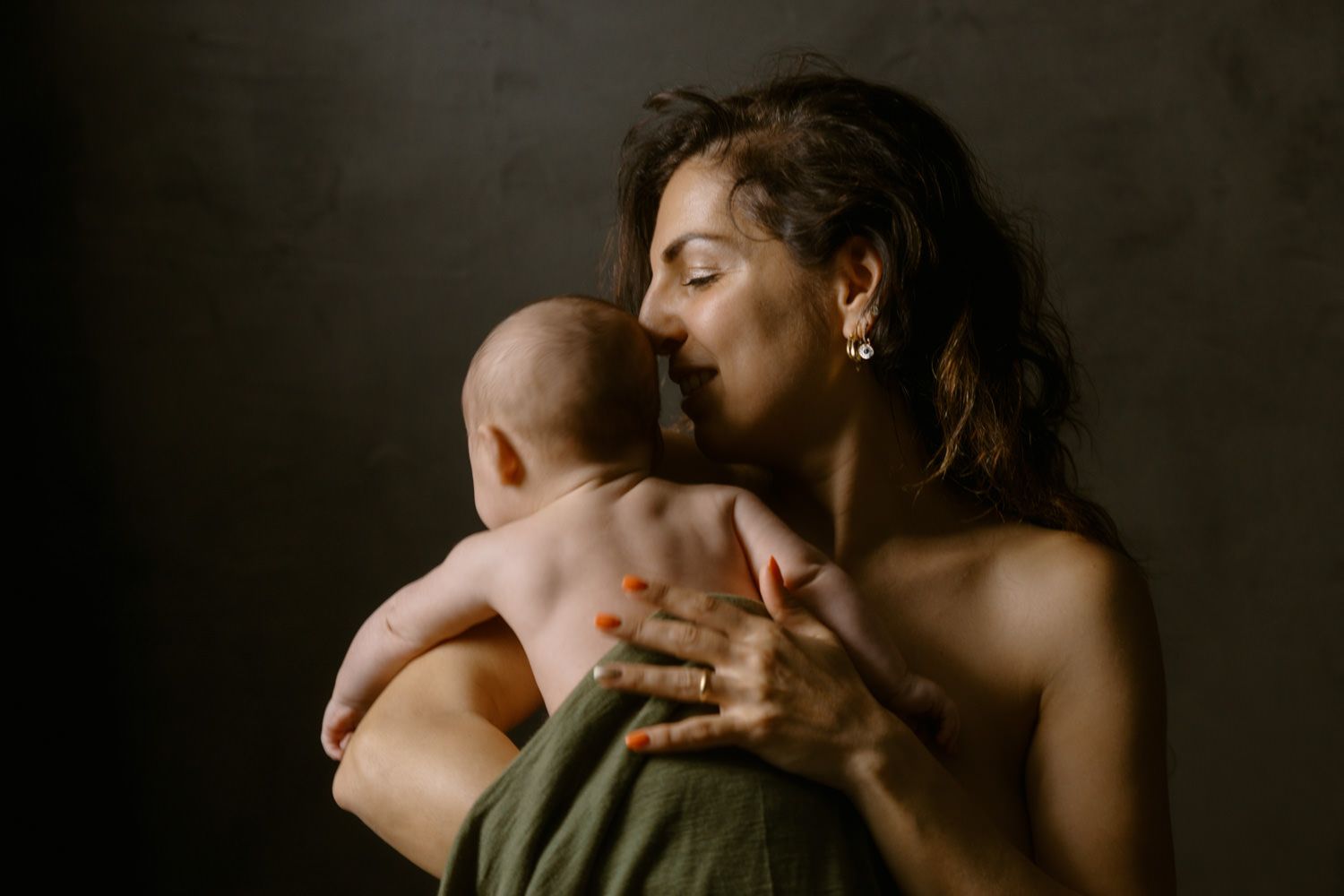 A mother tenderly holds her newborn baby against her bare shoulder in a softly lit, moody portrait.