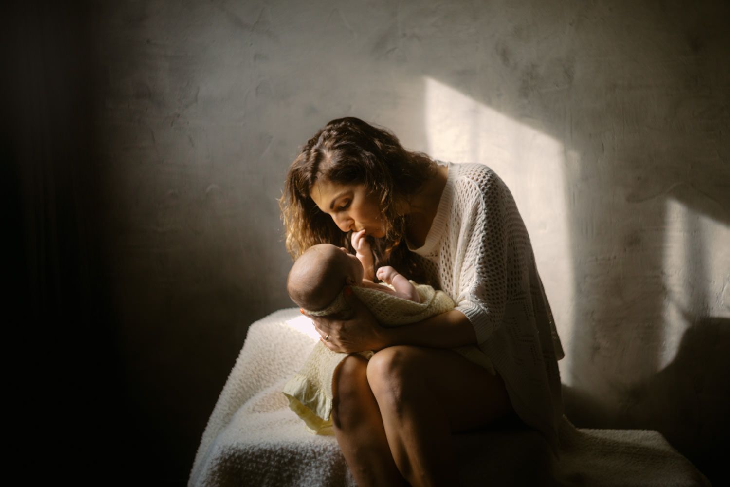 A mother tenderly holds and kisses her newborn baby in soft natural light.