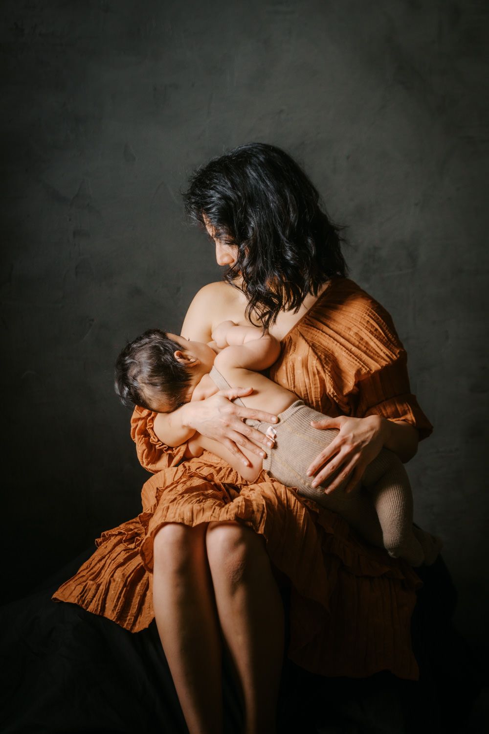 A mother in an orange dress tenderly breastfeeds her baby against a dark studio background.