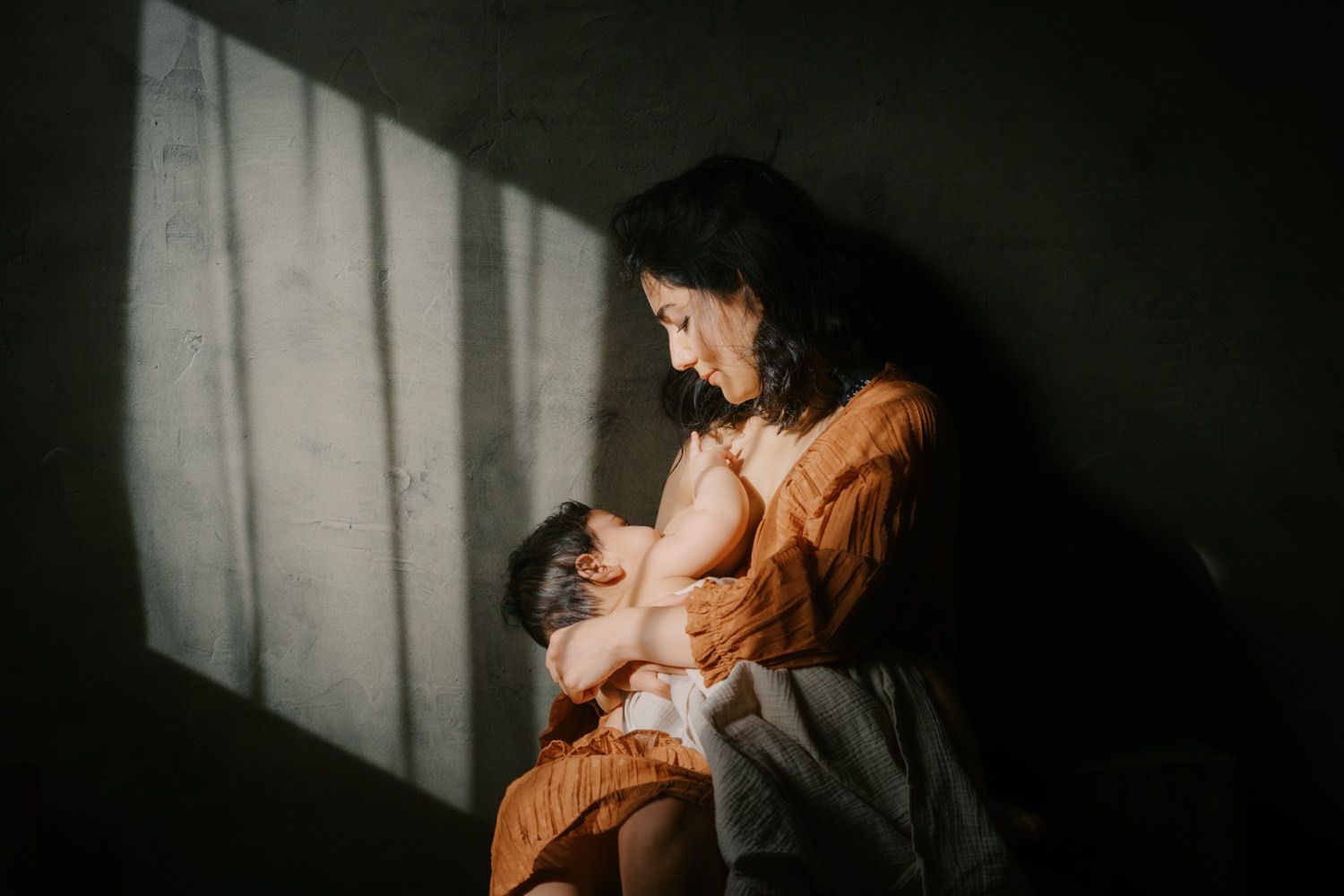 A mother tenderly breastfeeds her newborn in dramatic natural light against a textured dark wall.