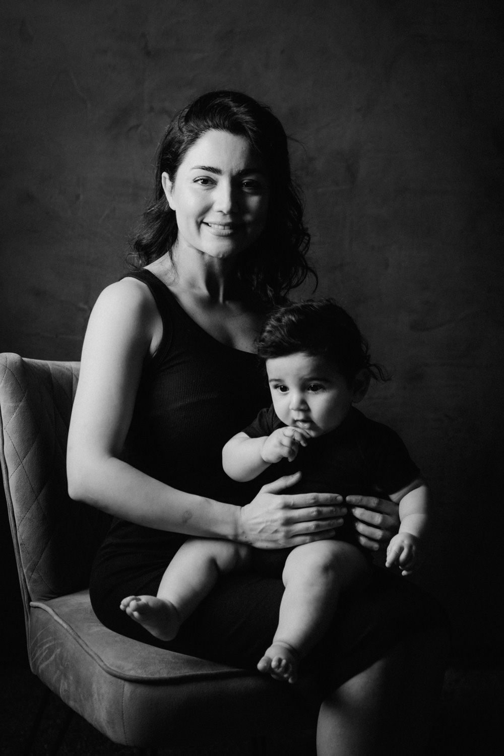 Black and white portrait of a smiling woman in black dress sitting in chair holding a cute baby on her lap.