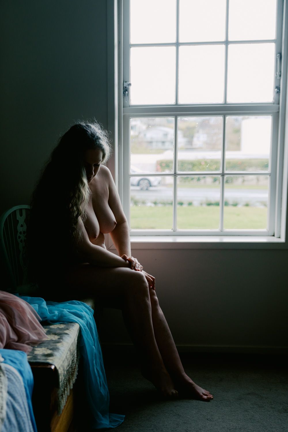 A nude woman sits on a bed by a large window, her long hair cascading down as she gazes outside.