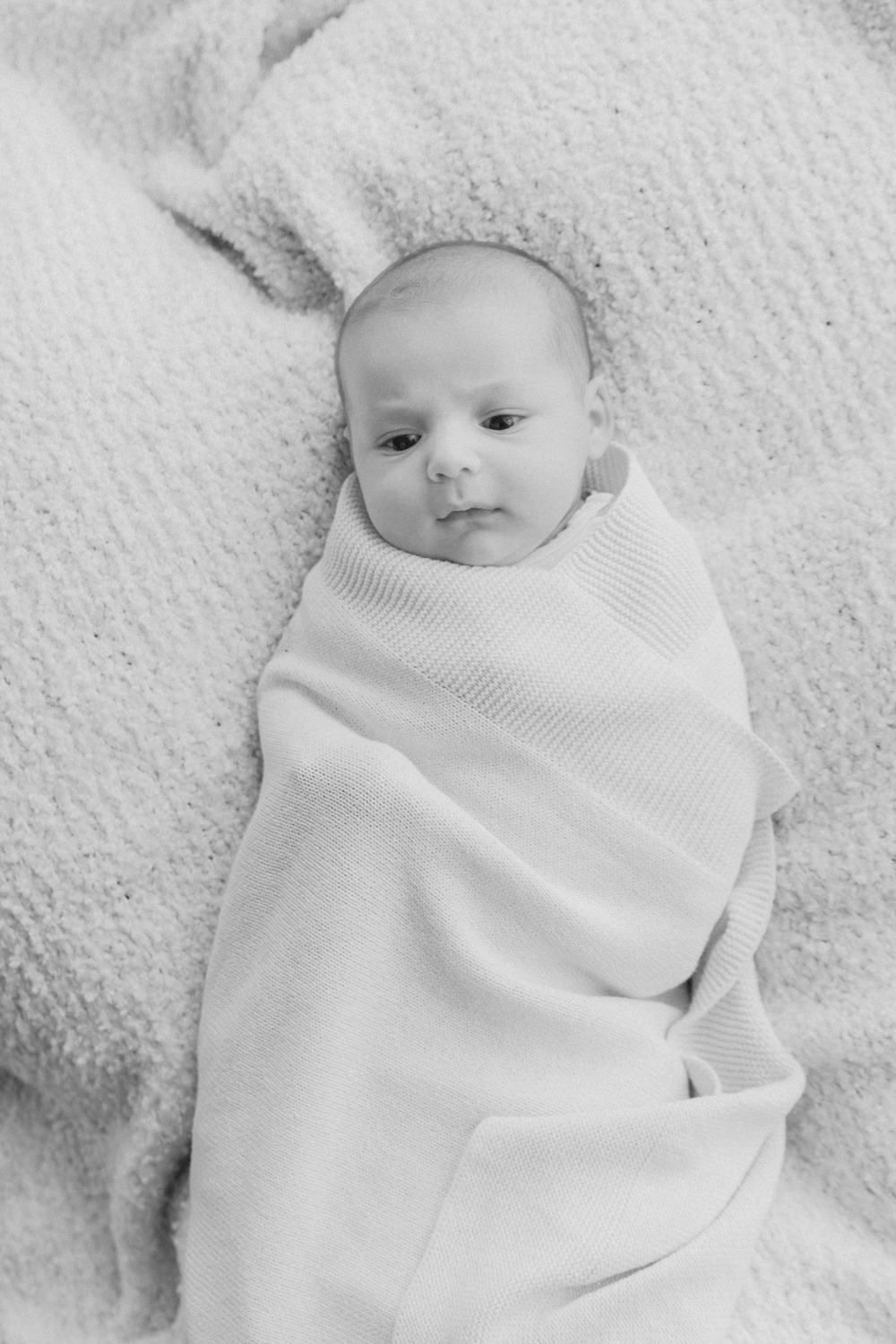 Black and white photo of a newborn baby swaddled in a soft white blanket, lying on a cozy textured surface.