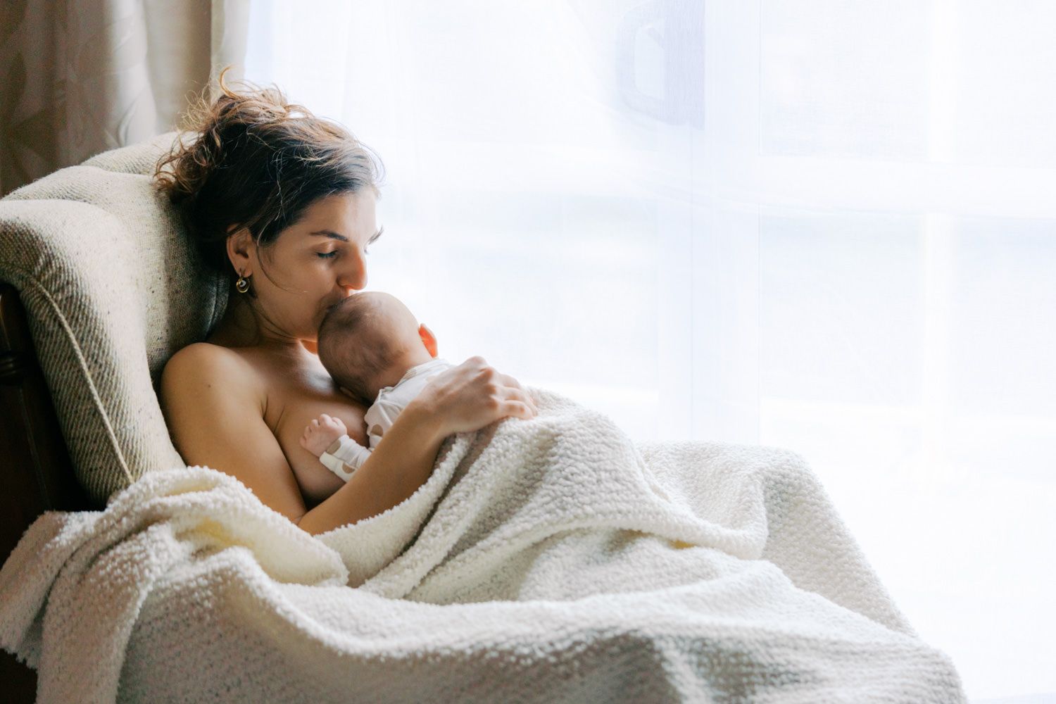 Mother tenderly kisses her newborn baby while resting in a chair wrapped in a soft white blanket.