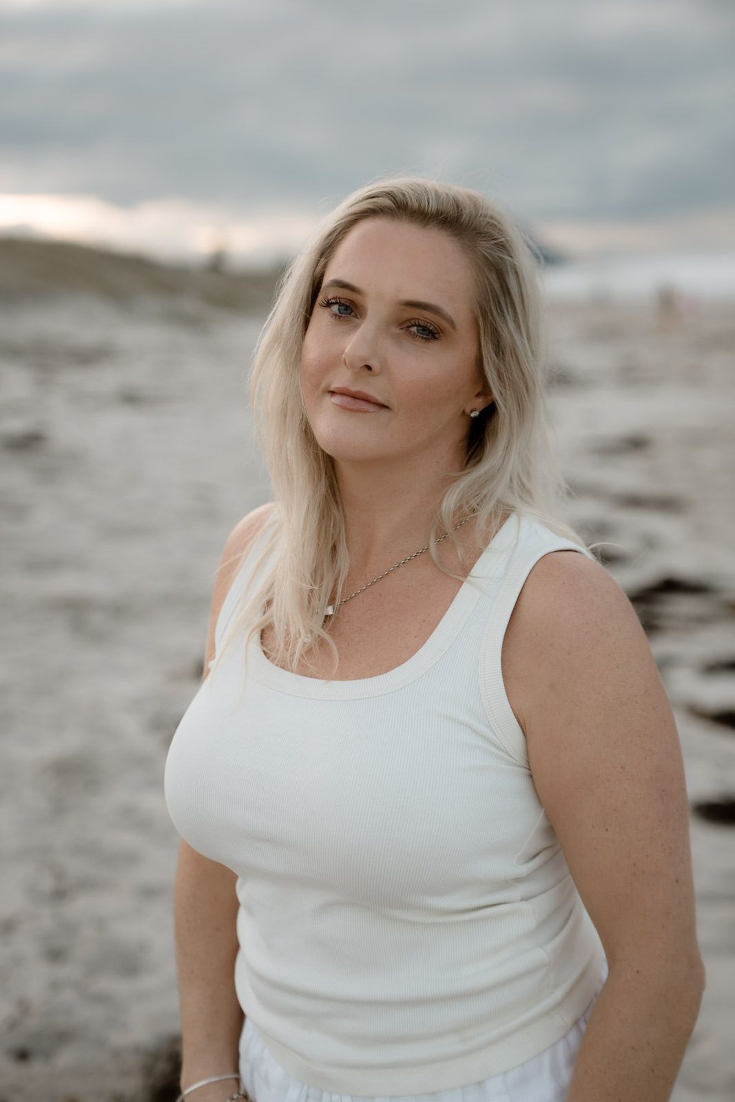 Blonde woman in white tank top posing on a sandy beach with cloudy sky background.