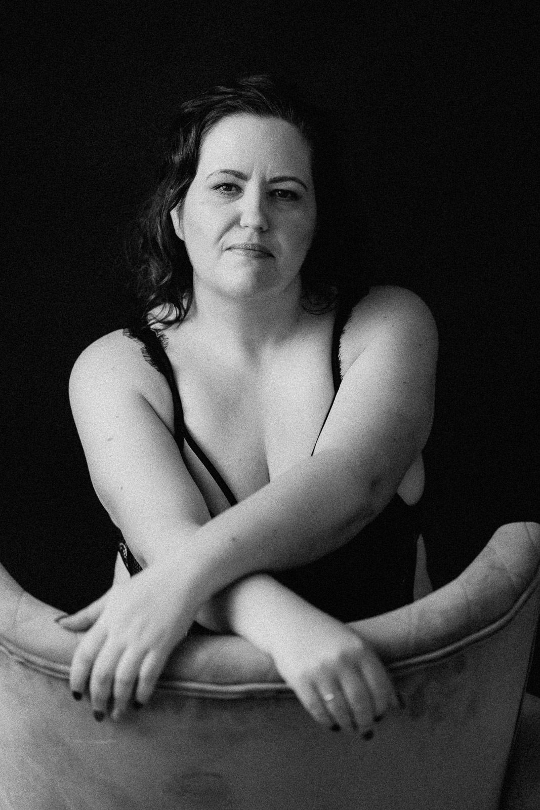 Black and white portrait of a woman leaning on a chair back, arms crossed, against a dark background.