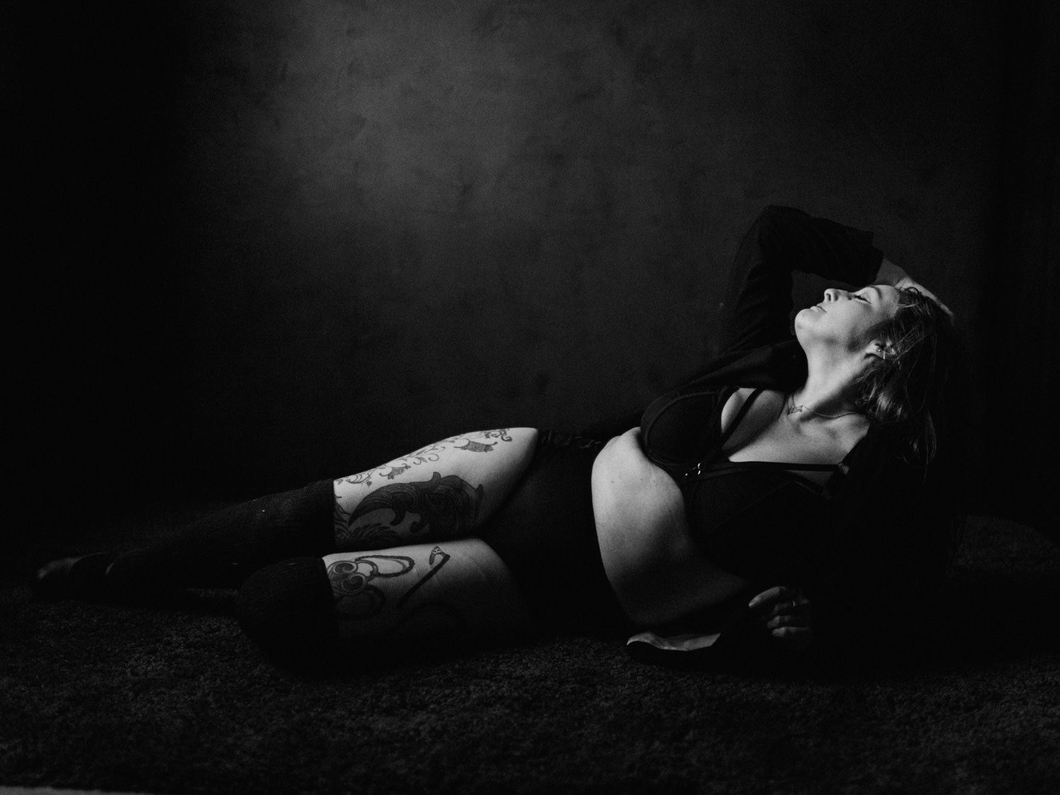 Black and white photo of a tattooed woman lying on the floor in dark clothing, looking upward dramatically.