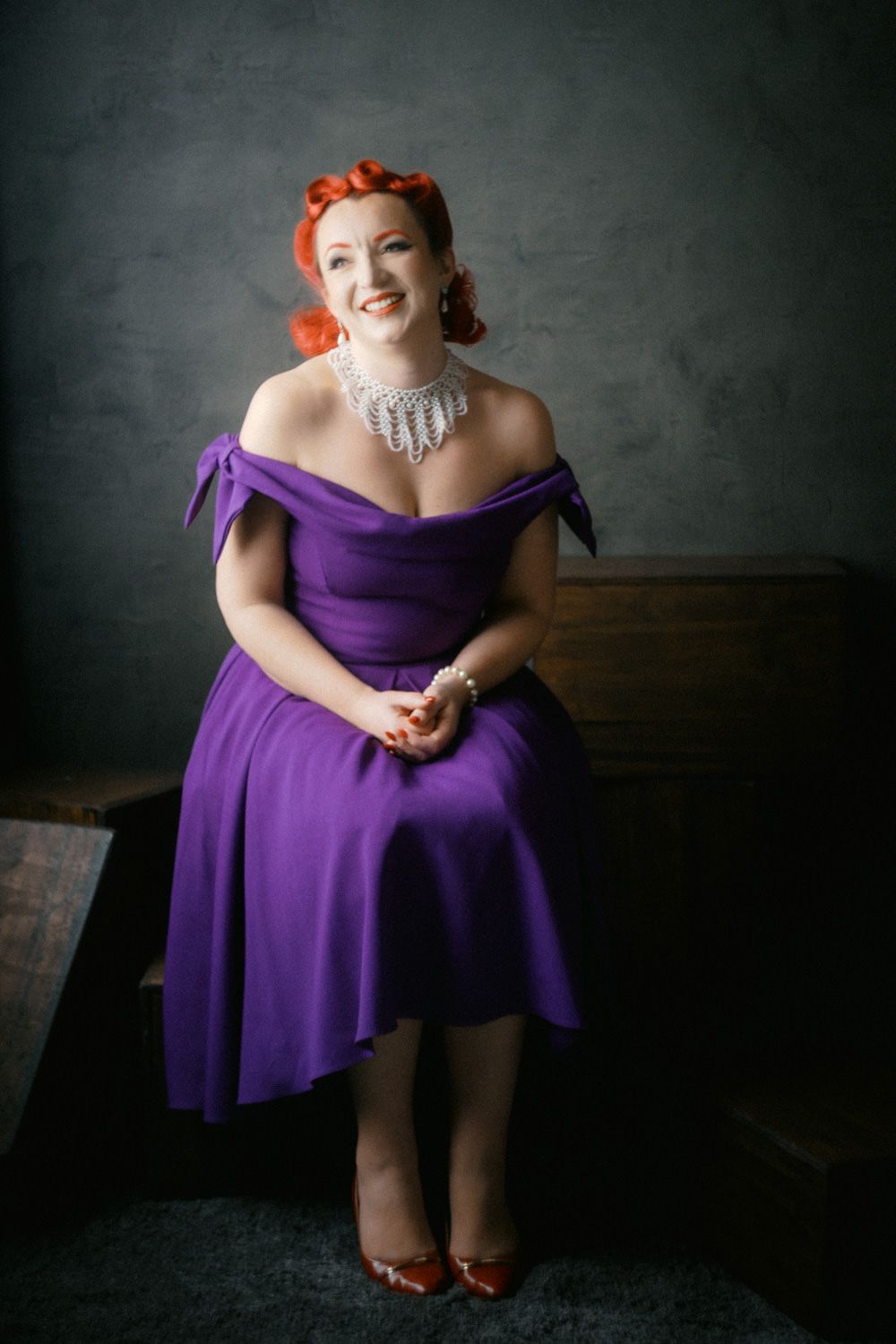 Woman in purple off-shoulder dress with red hair and pearl necklace smiling while seated in vintage-style portrait.