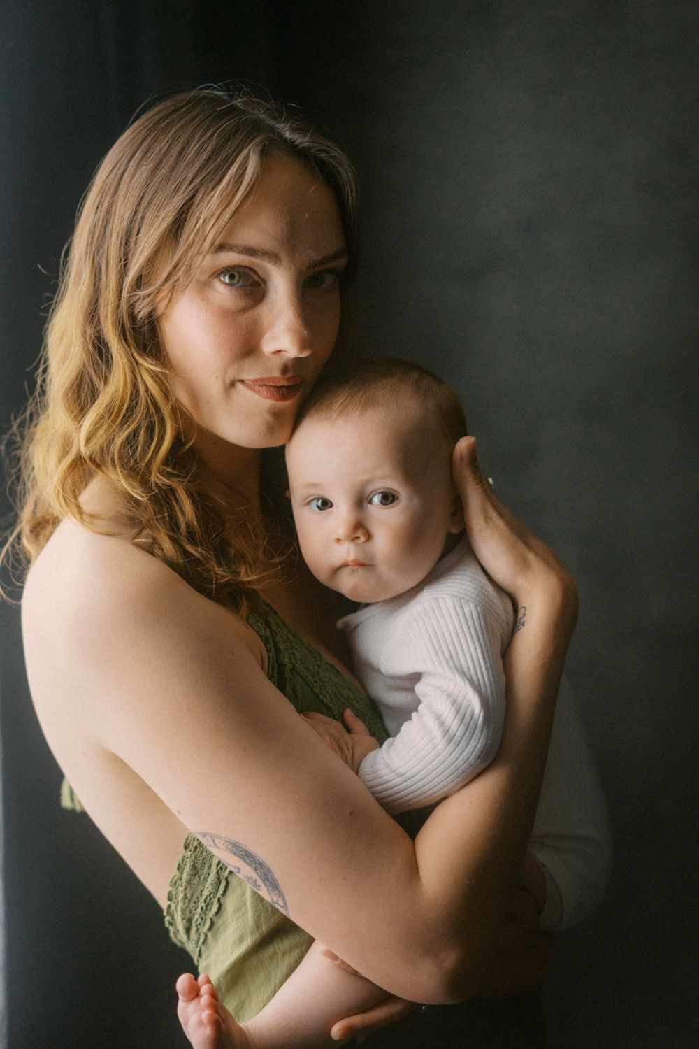 Young mother with wavy hair tenderly holds her infant baby in white onesie against dark background.