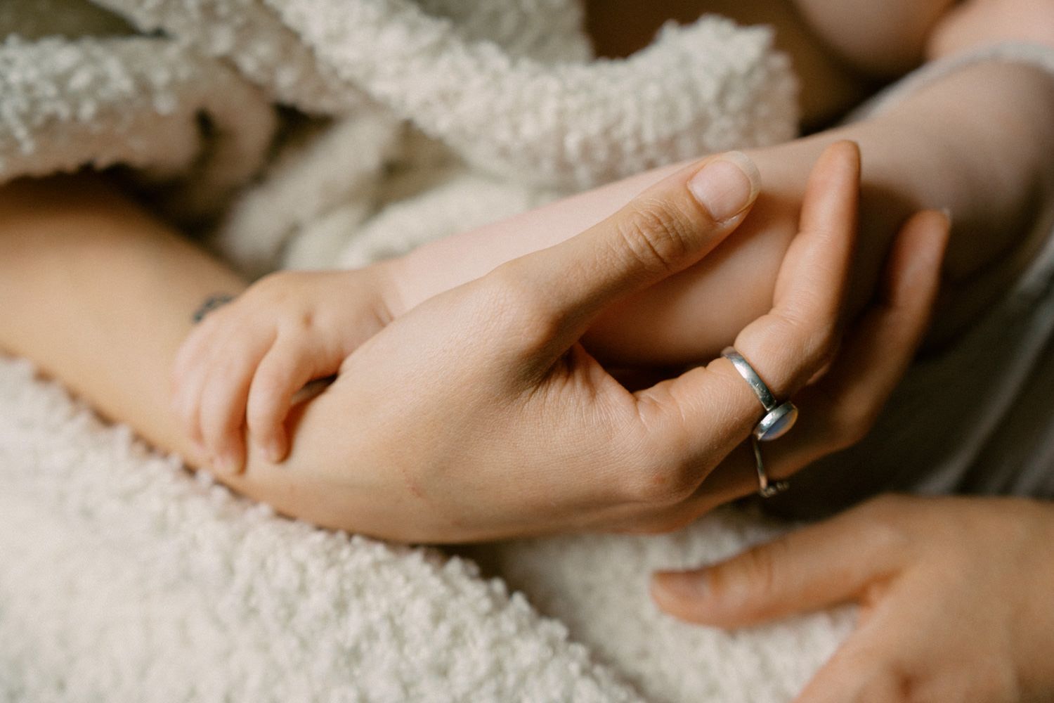 A mother gently holds her newborn baby's tiny hand, wearing a delicate silver ring, on a soft white blanket.