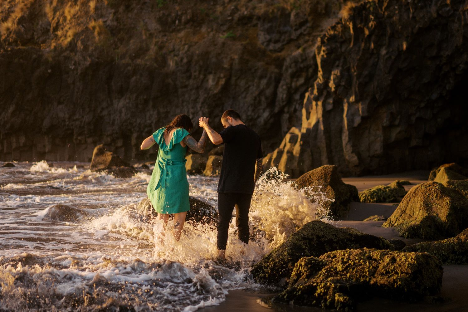 Couple in green dress and black outfit standing in ocean waves near rocky cliffs during golden hour.