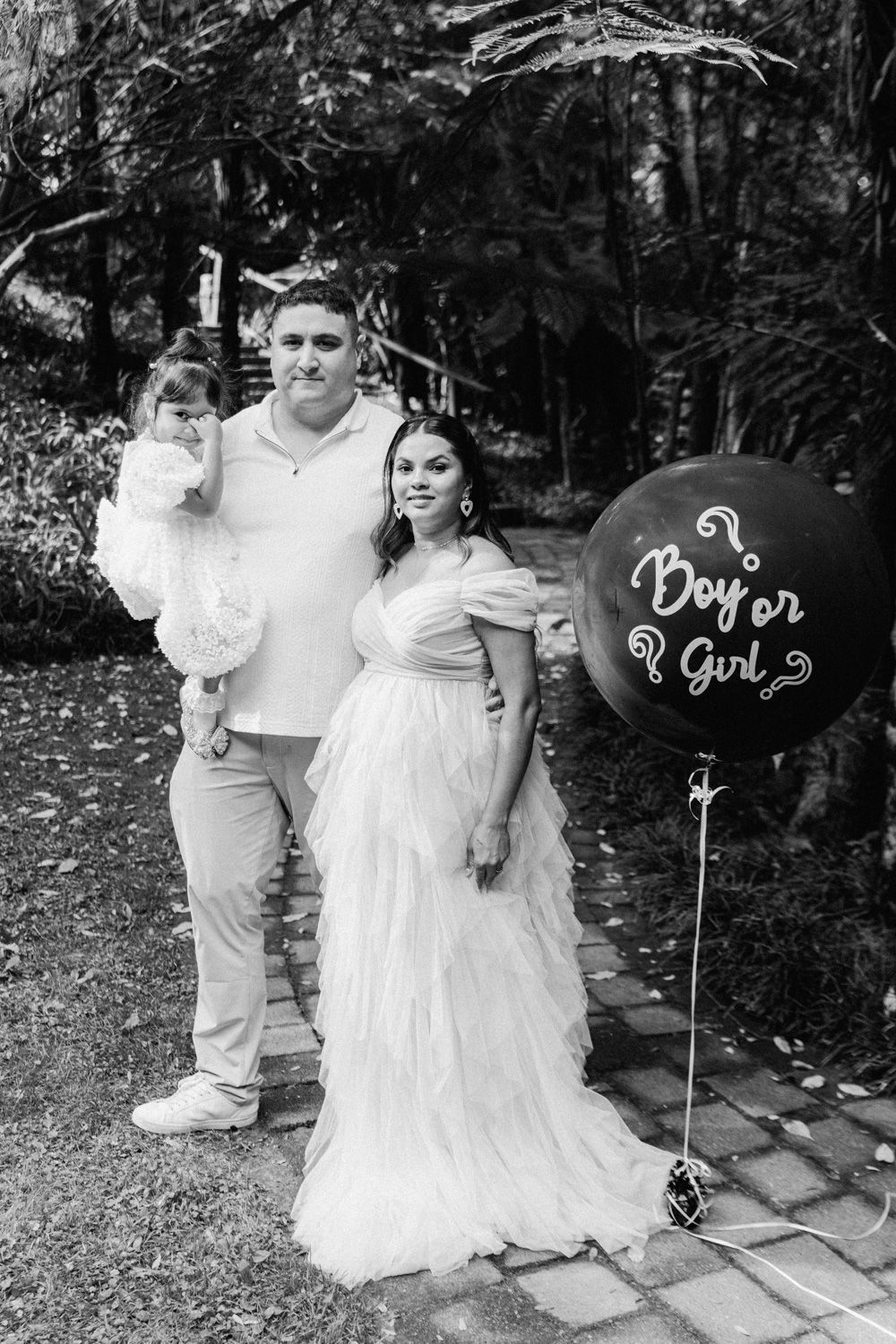 Black and white gender reveal photo of pregnant couple holding toddler with Boy or Girl balloon outdoors.