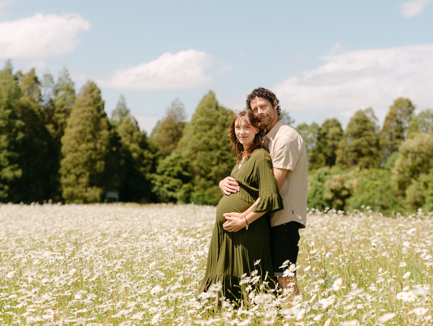 Couple poses in a daisy field, woman in green dress cradling baby bump, surrounded by trees under blue sky.
