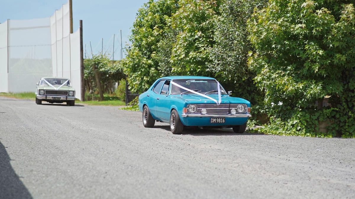 A vintage blue car decorated with white ribbons leads a wedding procession down a quiet rural road.