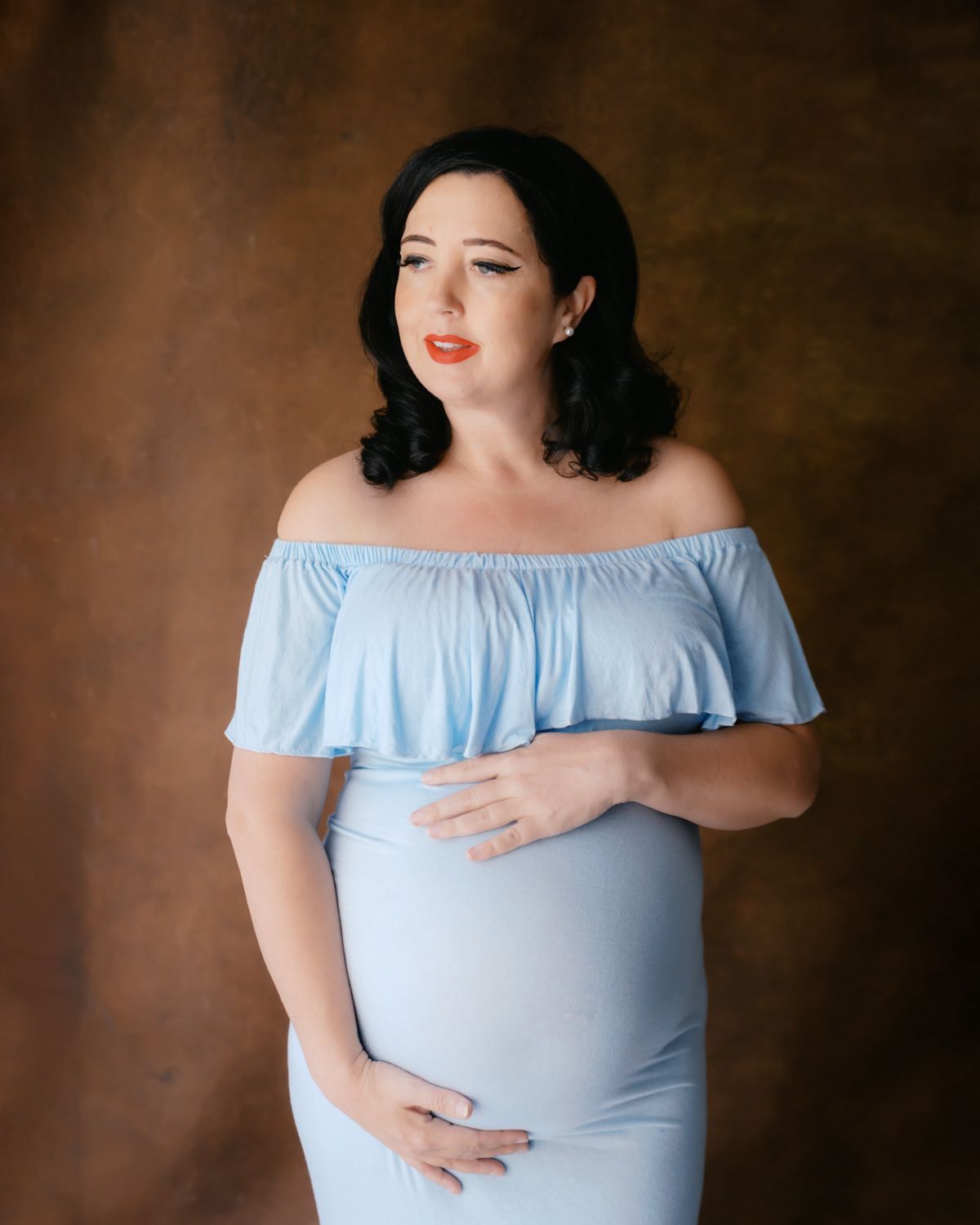 Pregnant woman in light blue off-shoulder maternity gown cradling her baby bump against a brown studio backdrop.