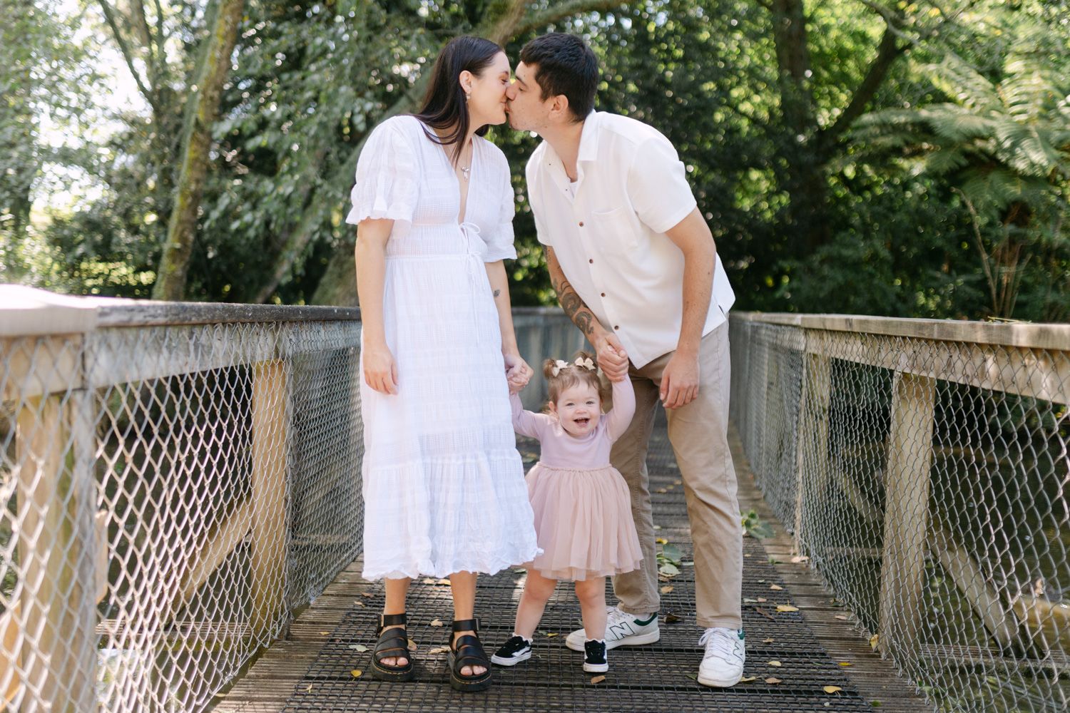 A happy family with parents kissing and toddler girl in pink dress holding hands on a bridge in a lush green park.