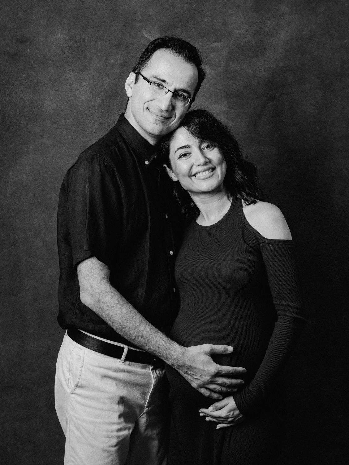 Black and white maternity photo of a smiling couple, man embracing pregnant woman from behind in a studio.