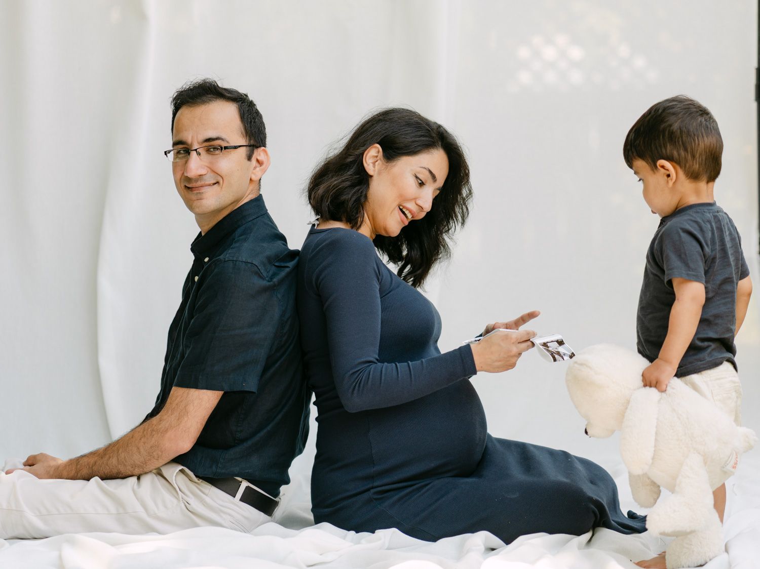 Pregnant woman sitting back-to-back with husband while toddler plays beside them during maternity photo session.
