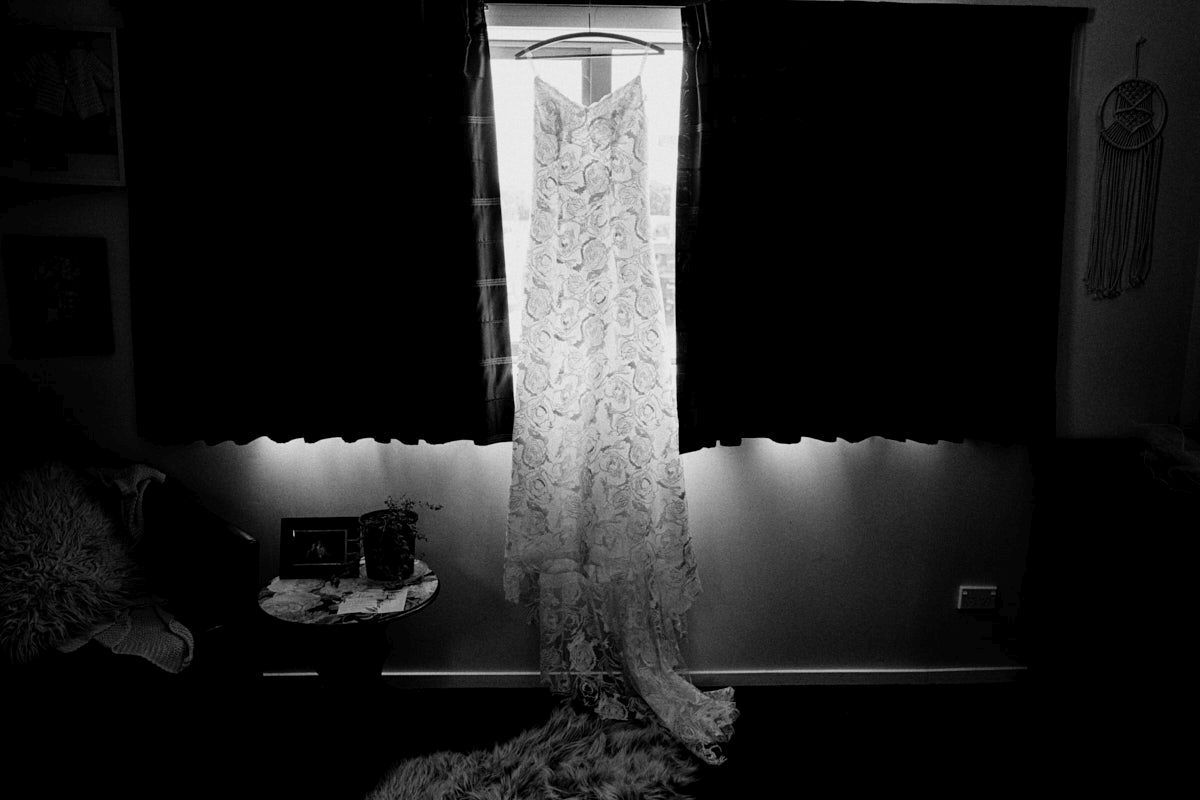 Black and white photo of an elegant lace wedding dress hanging against a bright window in a dimly lit room.