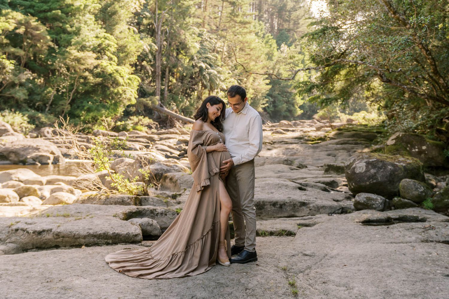 Couple poses on rocky riverbank surrounded by lush forest, woman in flowing tan gown with long train.