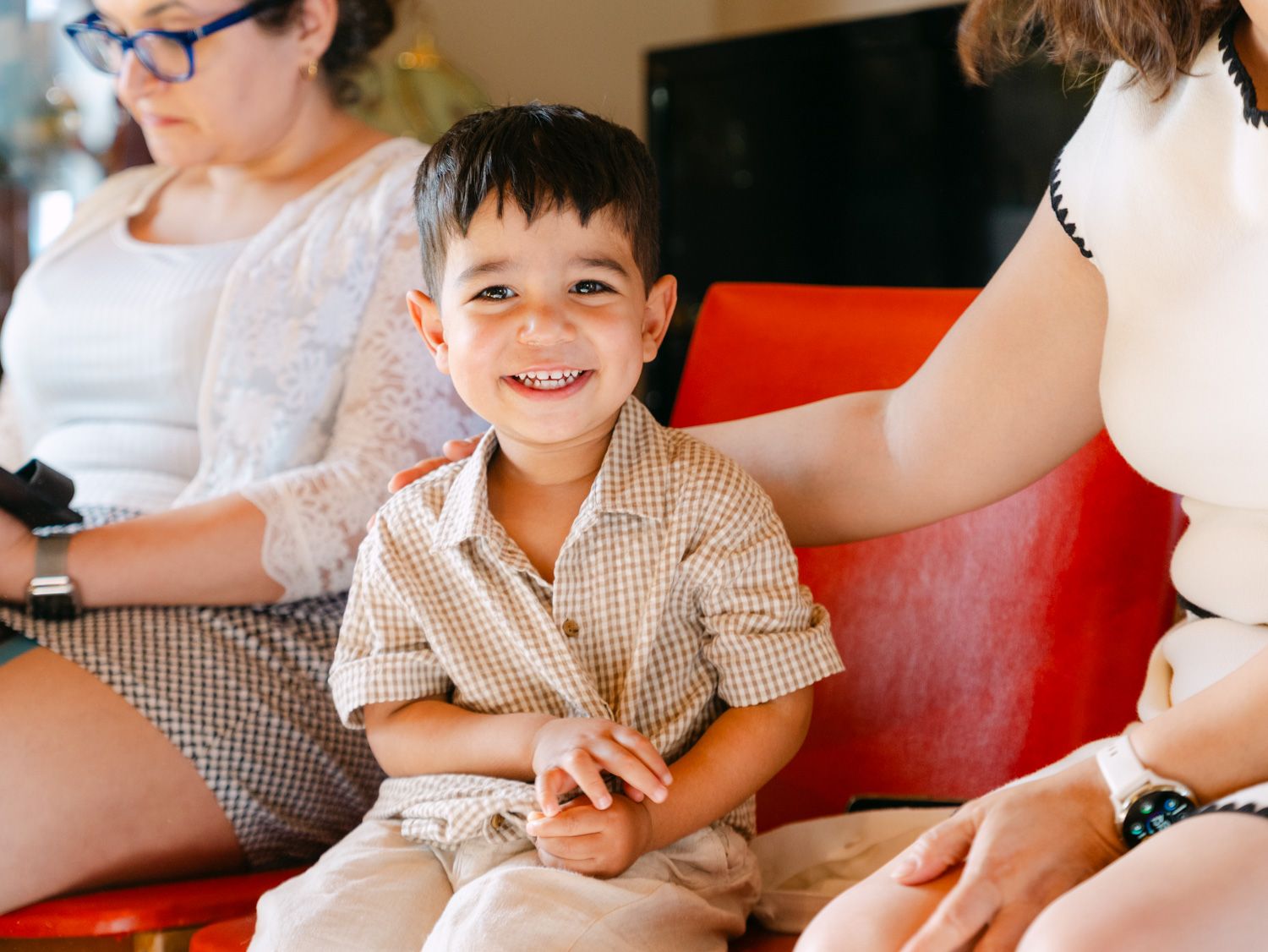 A smiling young boy in a checkered shirt sits happily between two women on a red chair.