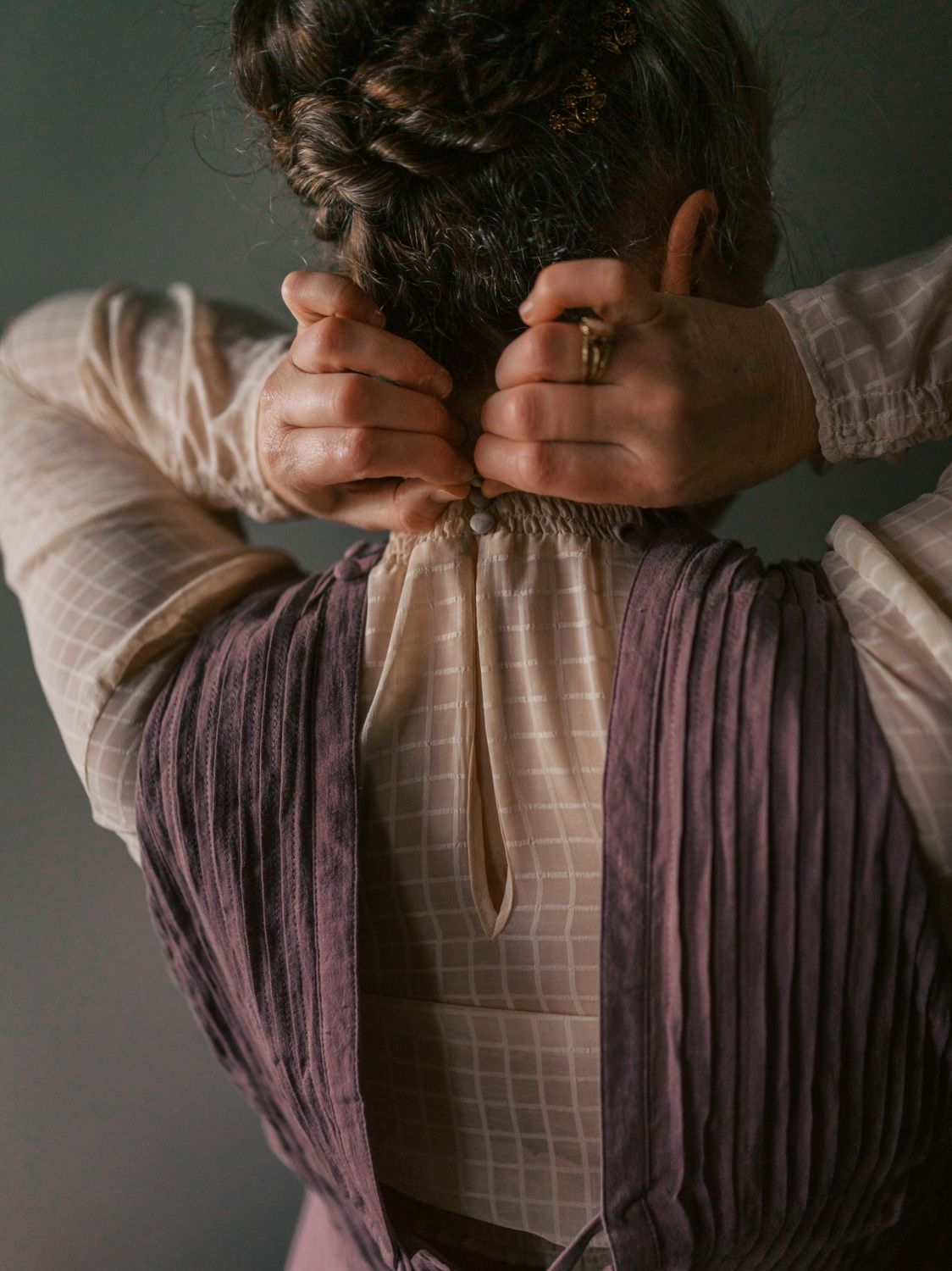 Woman in vintage purple and cream grid blouse fastening the back neckline with both hands, rear view.