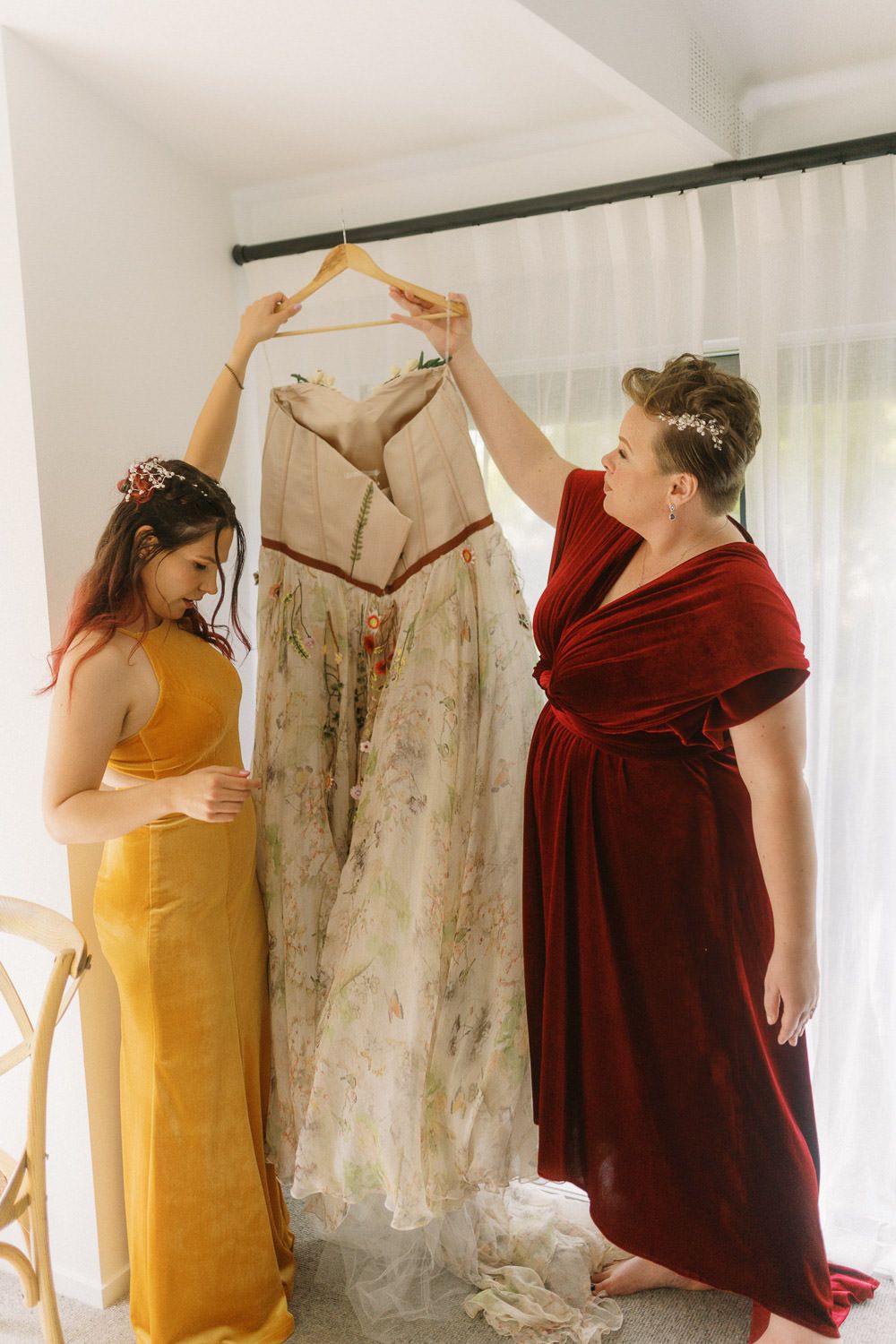 Two bridesmaids in red velvet and yellow dresses admire a floral wedding gown on a hanger.