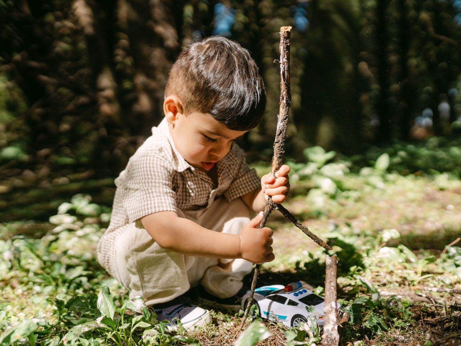 Young boy playing outdoors in forest, examining a stick while crouching on grass with a toy car nearby.