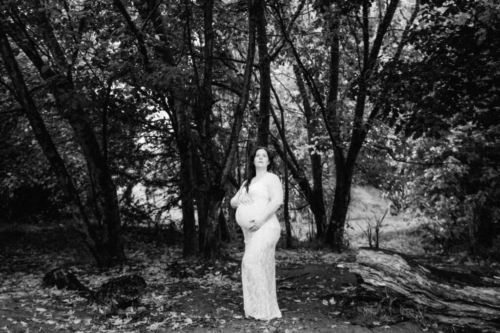 Black and white maternity photo of pregnant woman in lace gown standing among trees in a forest setting.