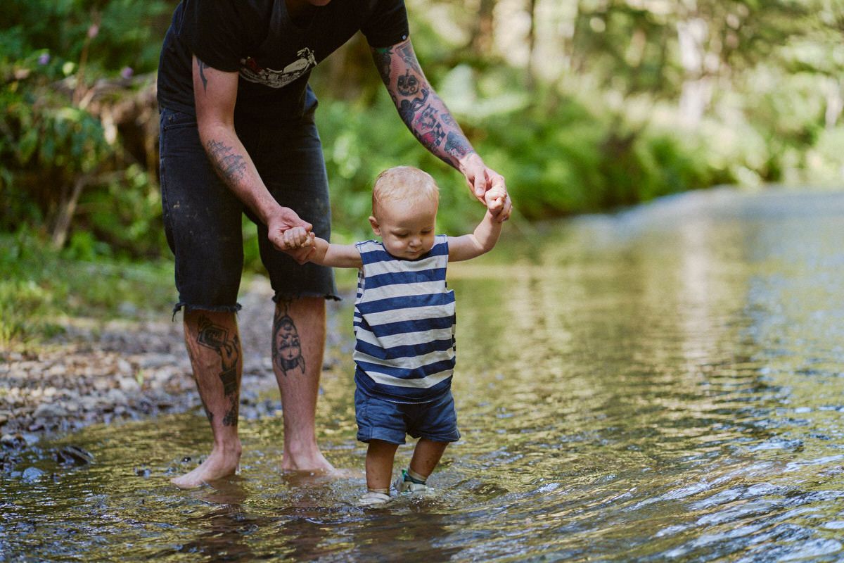 Tattooed father holding hands with toddler son wading in shallow creek surrounded by lush green forest.