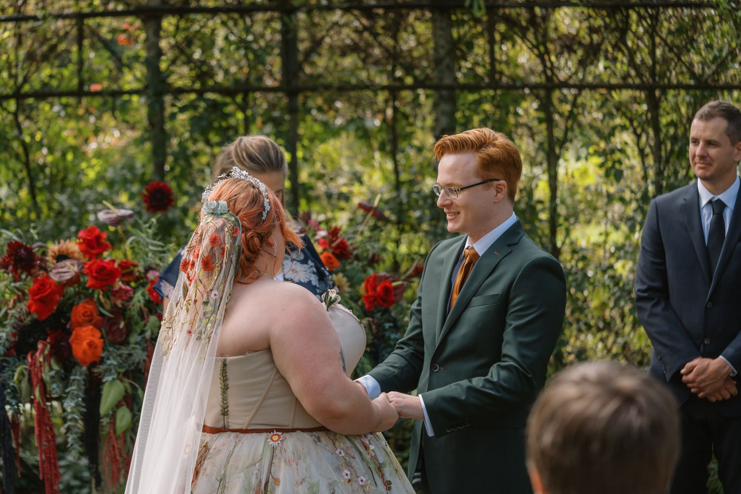 Bride and groom holding hands during outdoor garden wedding ceremony with red floral arrangements.