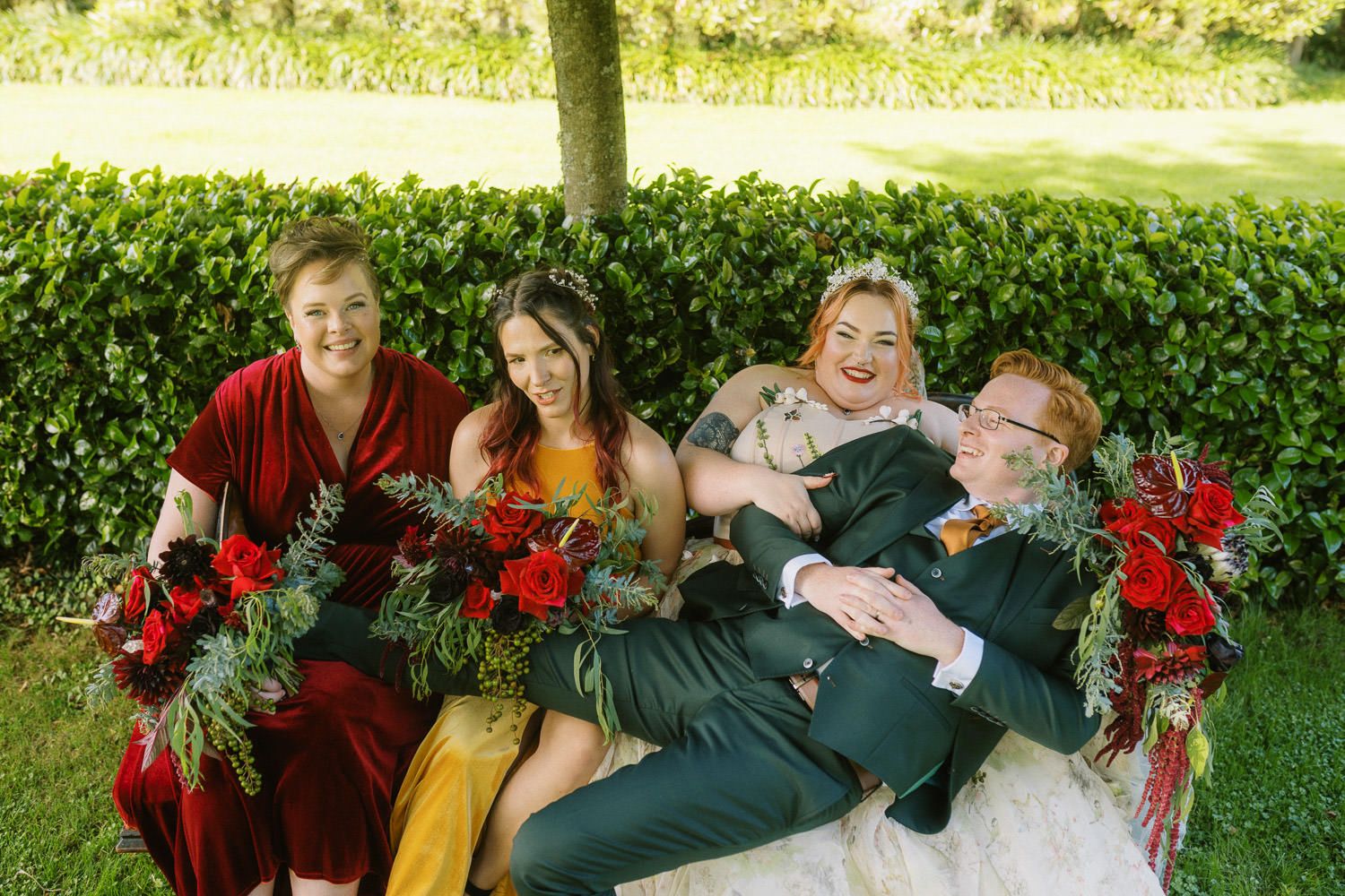 Bridal party laughing on bench with red bouquets, bridesmaids in burgundy and gold dresses, groom in teal suit.