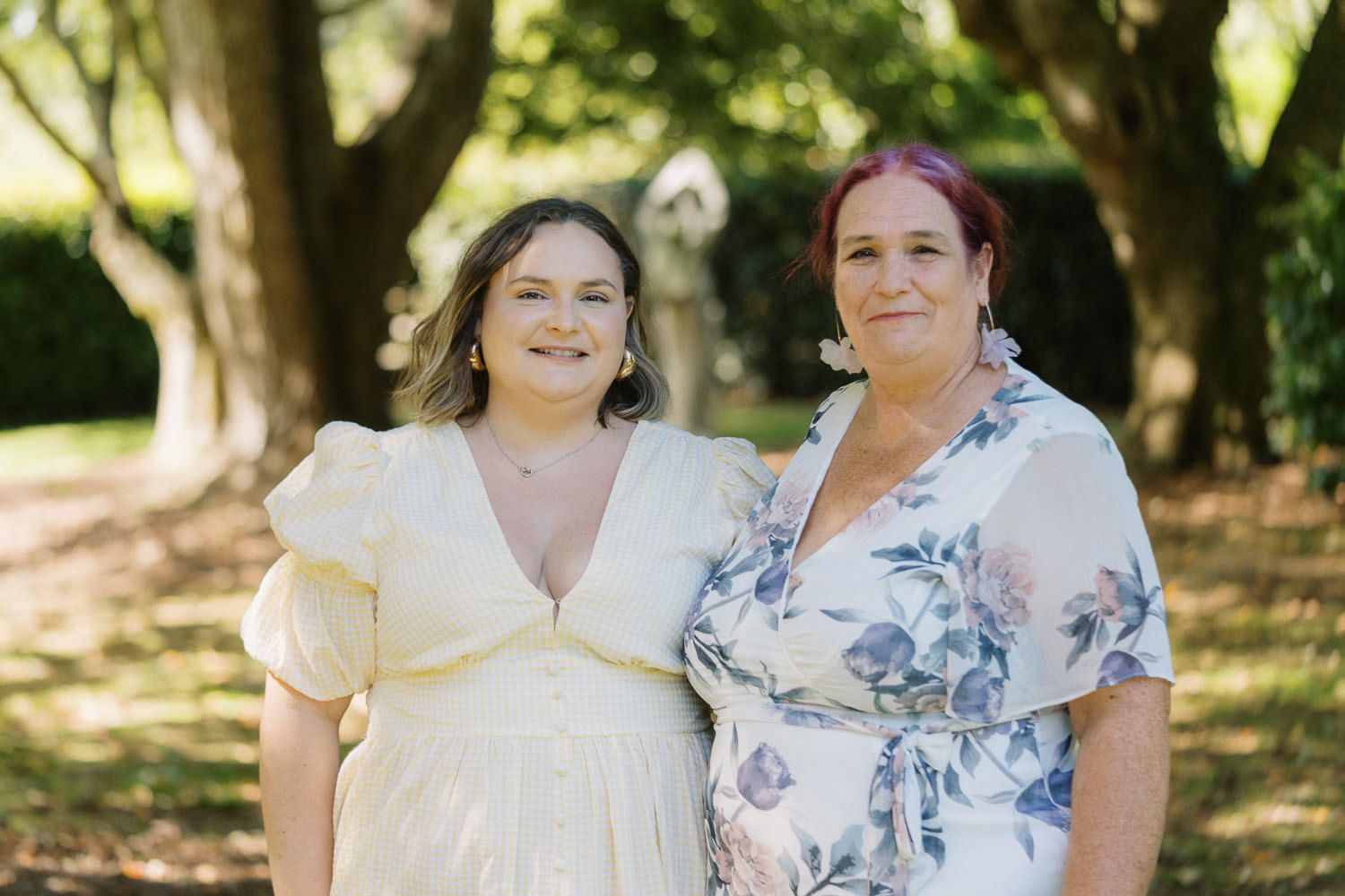 Two women smiling together outdoors in a lush green garden, wearing elegant dresses for a special occasion.