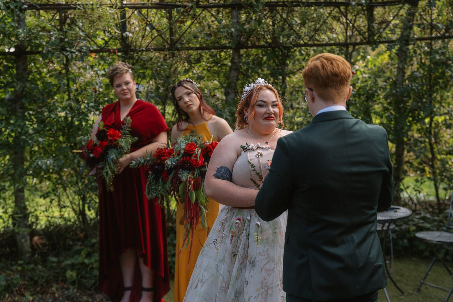 Bride in floral gown exchanges vows with groom outdoors, flanked by bridesmaids in red and yellow dresses holding bouquets.