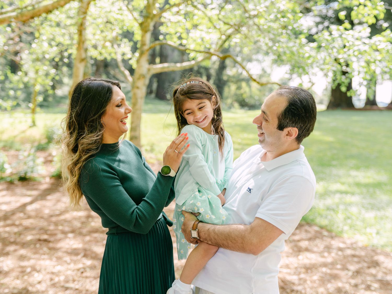 Family of three enjoying a sunny day in a lush green park, parents smiling at their young daughter.