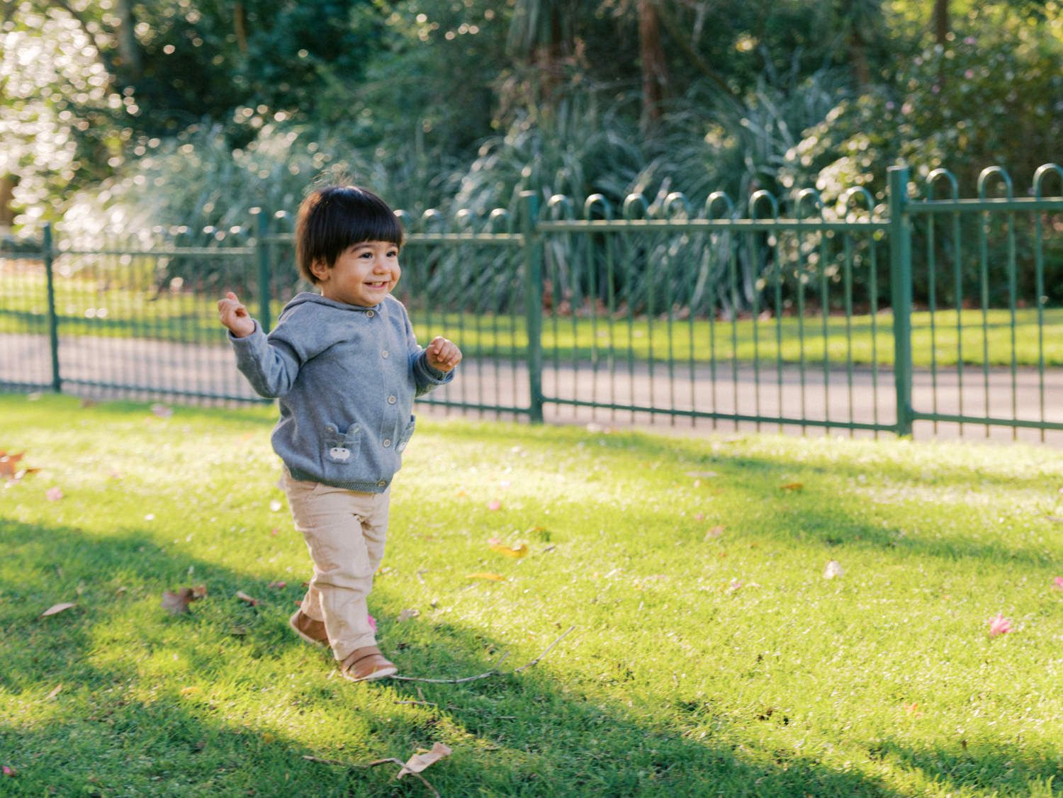 Toddler boy in gray hoodie and tan pants happily running on green grass in a sunny park with iron fence.