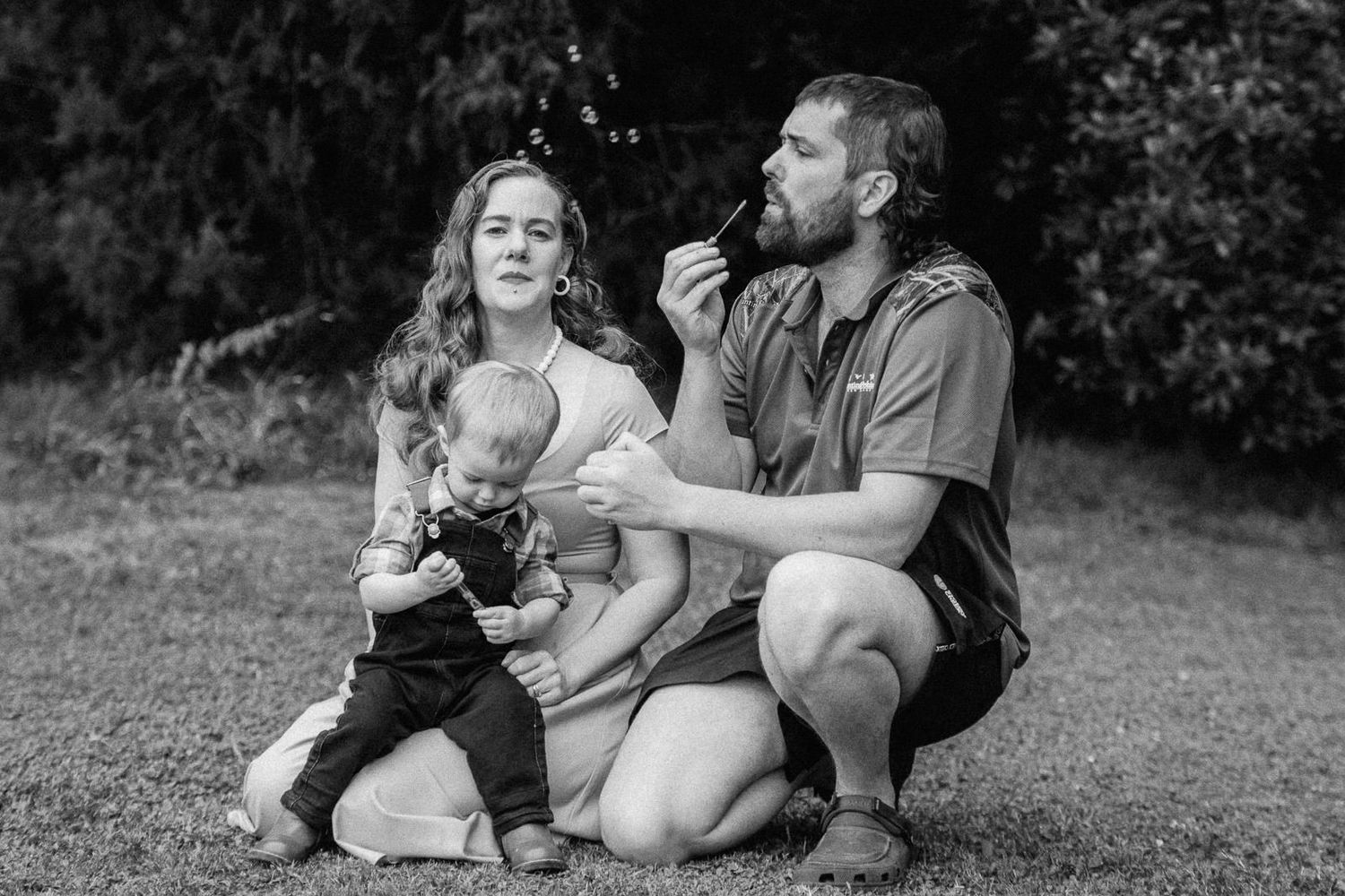 Black and white photo of a young family outdoors, mother holding baby while father blows bubbles on a grassy lawn.