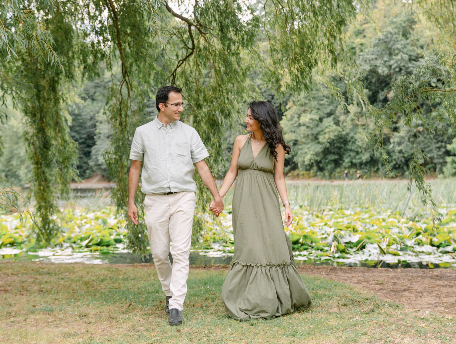 Couple holding hands walking through lush green park with willow trees, woman in olive maxi dress, man in casual attire.