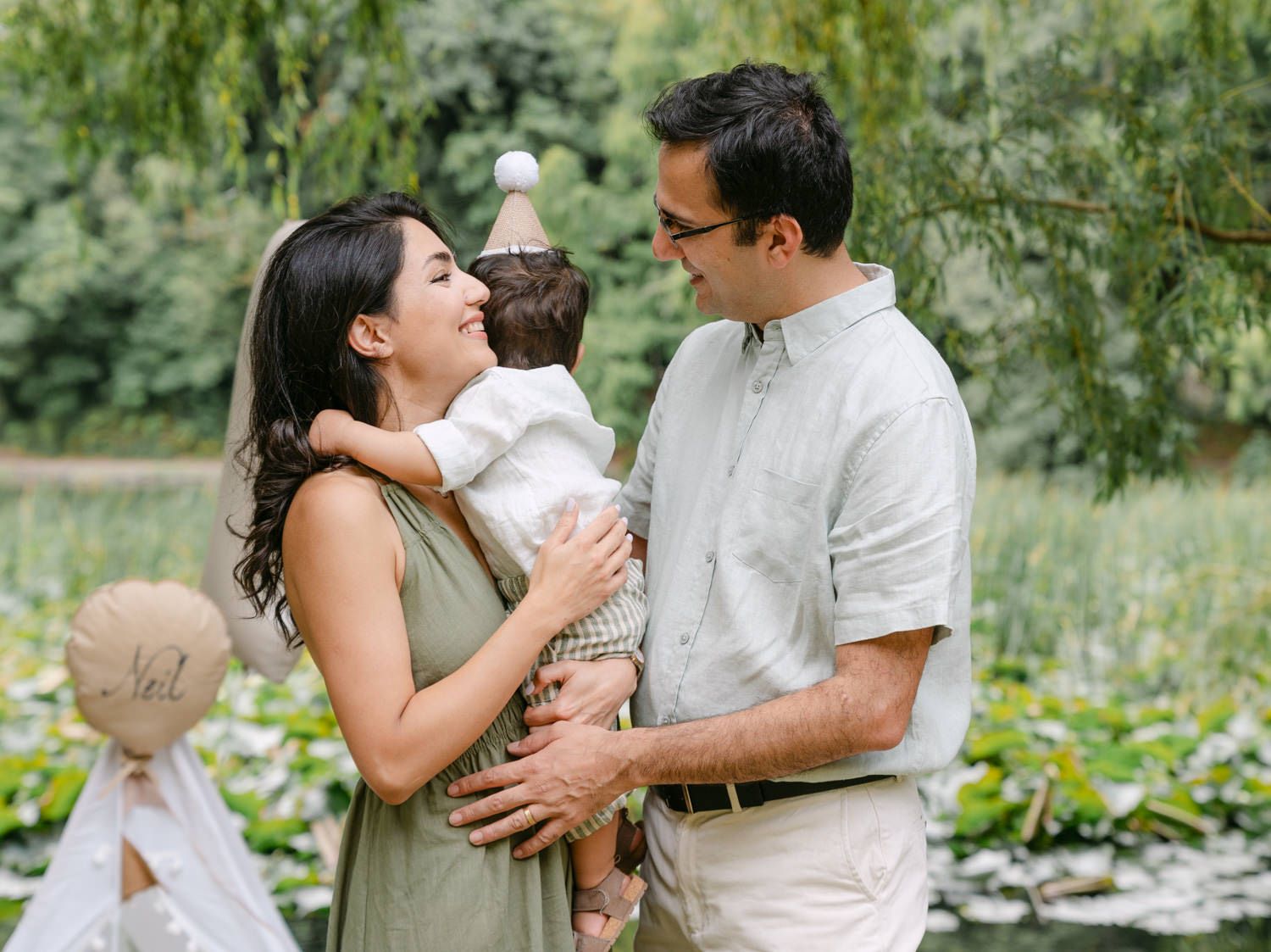 Happy couple smiling at their baby wearing a party hat outdoors near a pond surrounded by lush greenery.