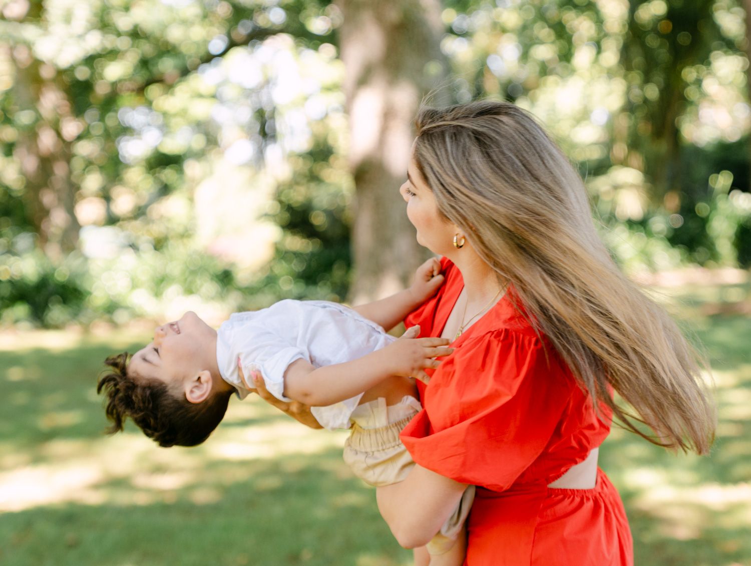 A mother in a red dress joyfully lifts her toddler outdoors in a lush green park setting.