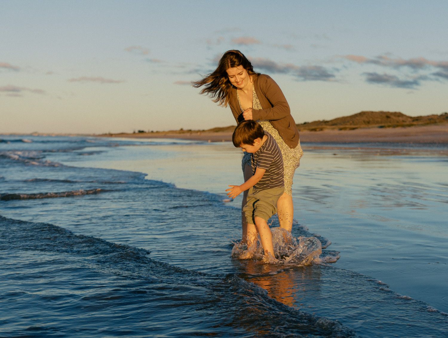 A mother and child laugh while splashing in ocean waves at sunset on a sandy beach.