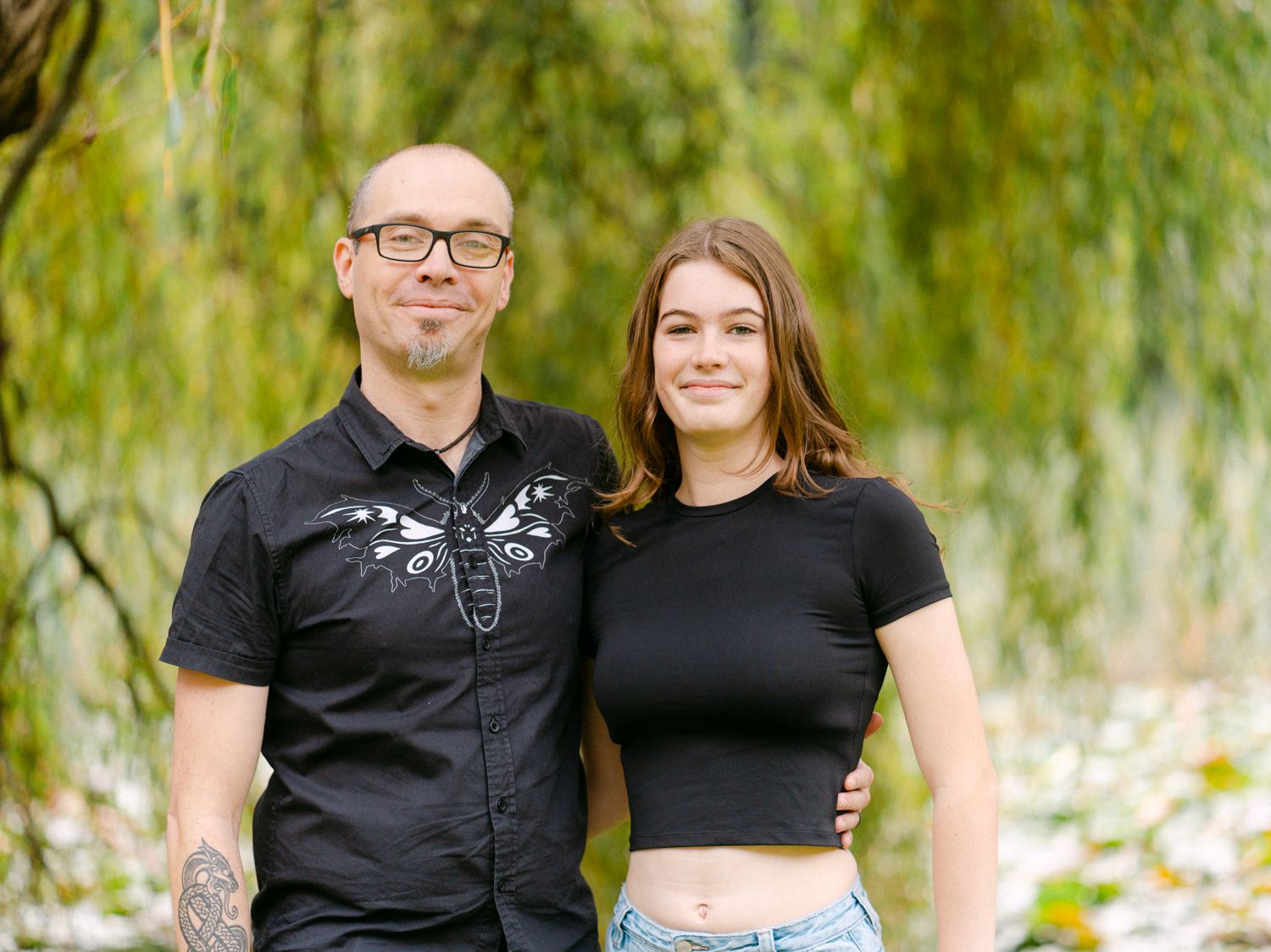 A bald man in a black shirt and a young woman in a black crop top pose together outdoors near a willow tree.