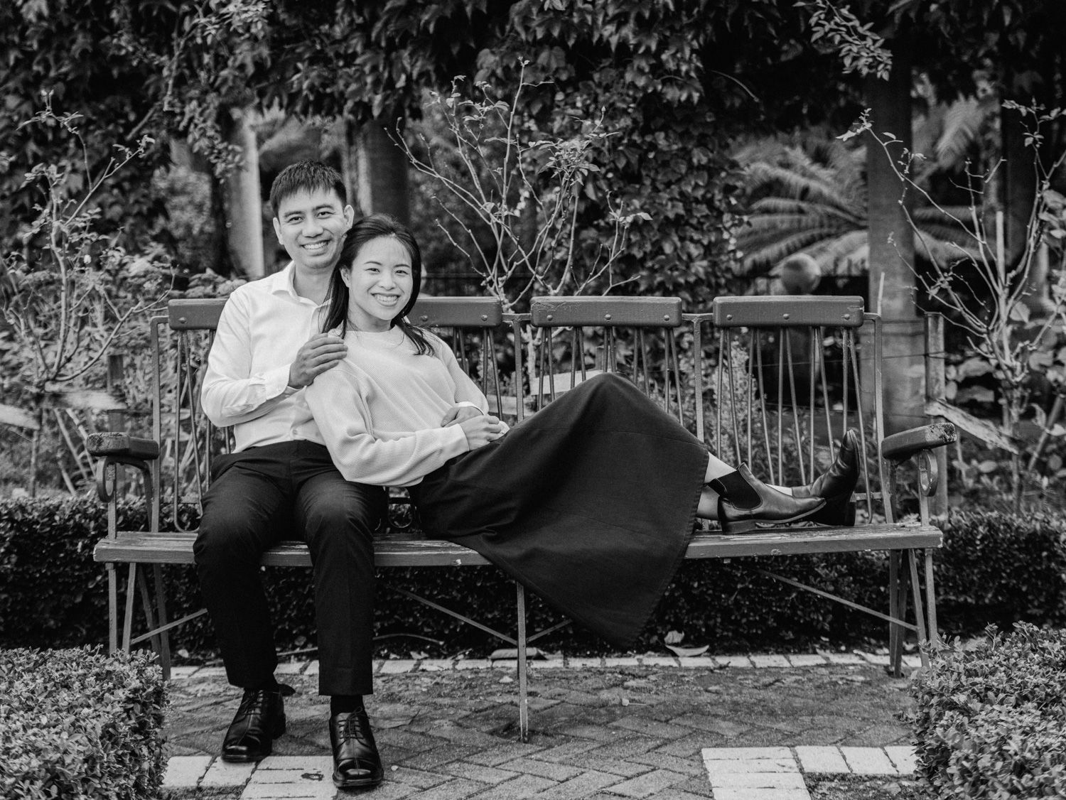 Couple sitting together on a wooden bench in a garden, smiling, black and white photo.